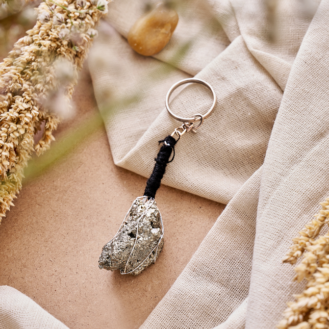 A lifestyle shot of a natural raw Peruvian Pyrite keychain, featuring a wire-wrapped metallic stone on a black cord, styled on a neutral fabric background with dried botanicals